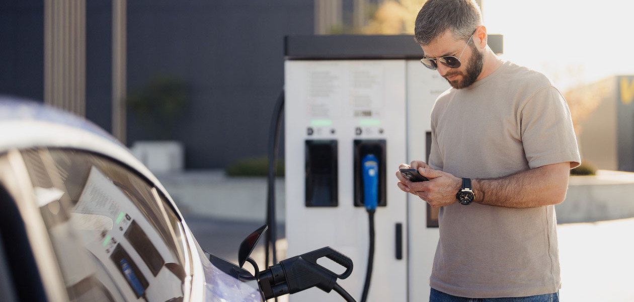 Homme qui regarde son téléphone portable devant une voiture et borne de rechargement