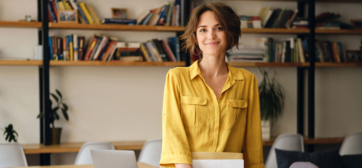 Young beautiful woman in yellow shirt leaning on desk with notepad and papers in hand while happily looking in camera in modern office