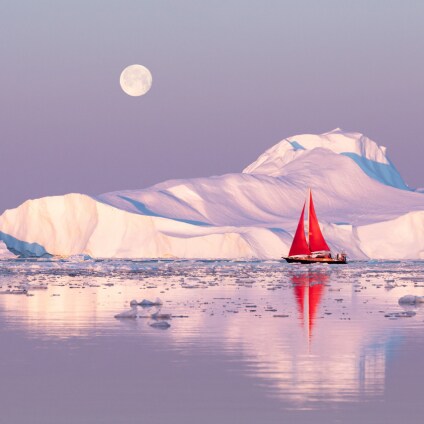 Little red sailboat cruising among floating icebergs in Disko Bay glacier during midnight sun season of polar summer. Ilulissat, Greenland.