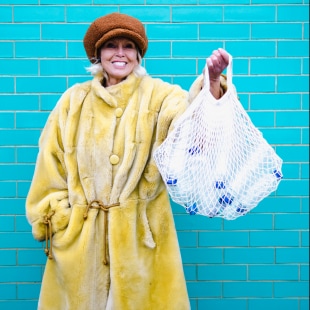 Femme senior souriante avec manteau jaune sur fond turquoise qui tient un sac filet rempli de bouteilles de platique
