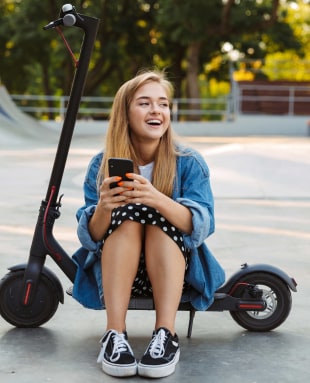 Happy teenage girl in park sit on scooter using mobile phone.