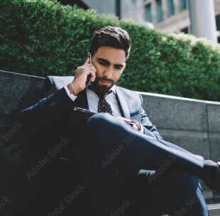 A young man in formal suit in the street talking on his mobile phone as looking at a report