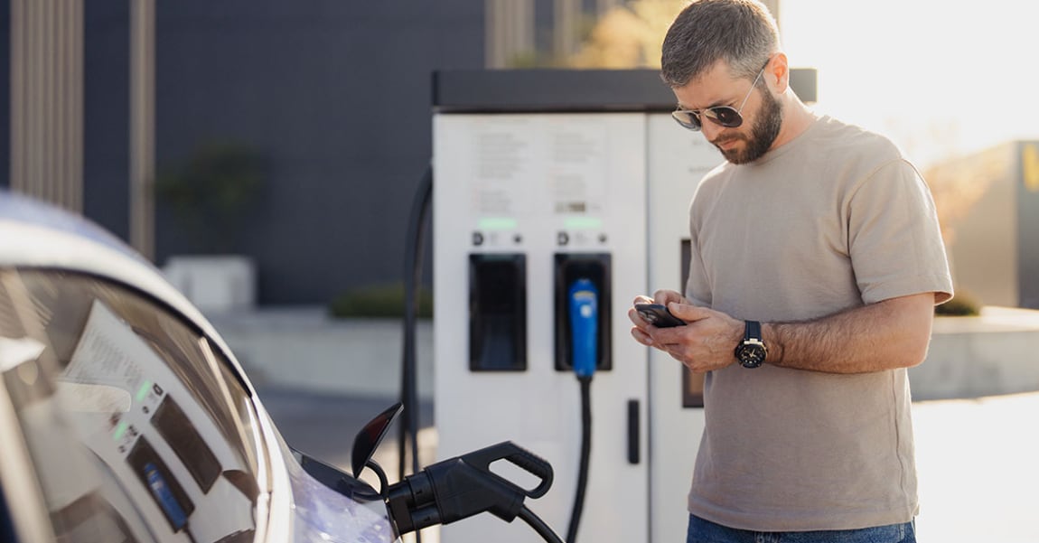 Man looks at cell phone standing in front of an electric vehicle that is charging