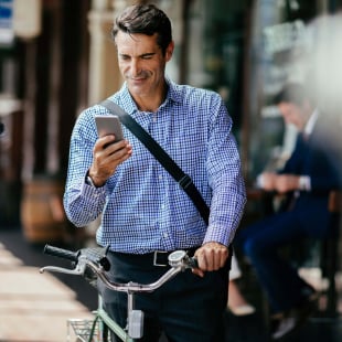 Middle aged businessman using a smartphone and pushing a bike through the city.