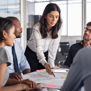 Managers around a table analysing documents. Woman standing points out figures with a pensil.